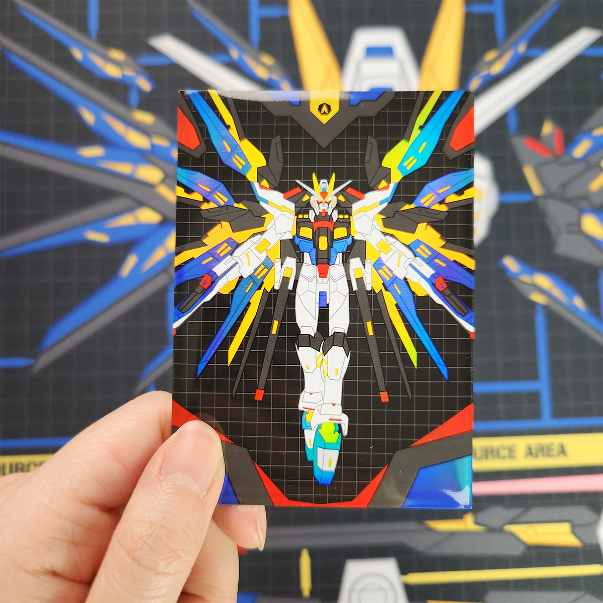 A hand holds a small printed card featuring a colorful robot illustration on a black grid background, with a blurred blue and yellow sci-fi scene behind it.