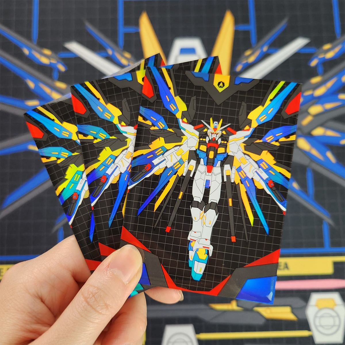 A hand holds three printed cards featuring a colorful mecha illustration on a black grid background, with a blurred blue and gray sci-fi themed backdrop behind.